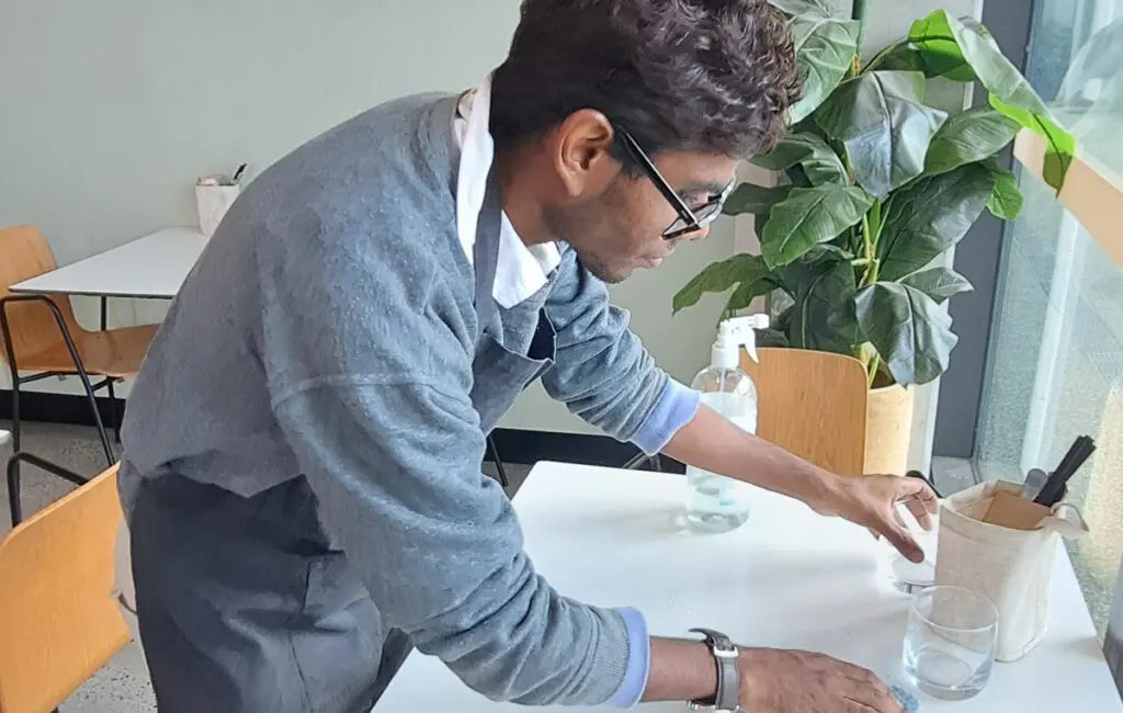 Man wearing glasses and an apron wipes a café table with a cloth near a spray bottle and glass.