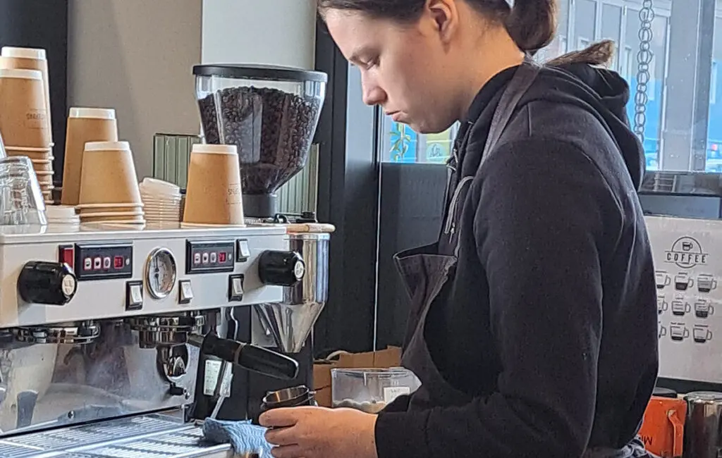 Barista in an apron steaming milk beside an espresso machine and coffee cups
