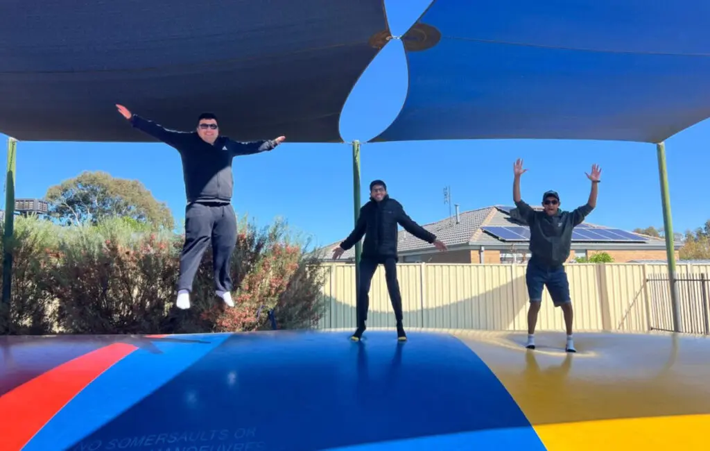 Three men jump with arms raised on a colourful outdoor trampoline under a shade sail.
