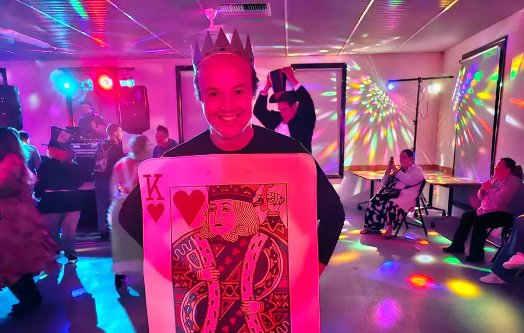 Man wearing crown and King of Hearts card costume smiles at a party with disco lights and guests dancing
