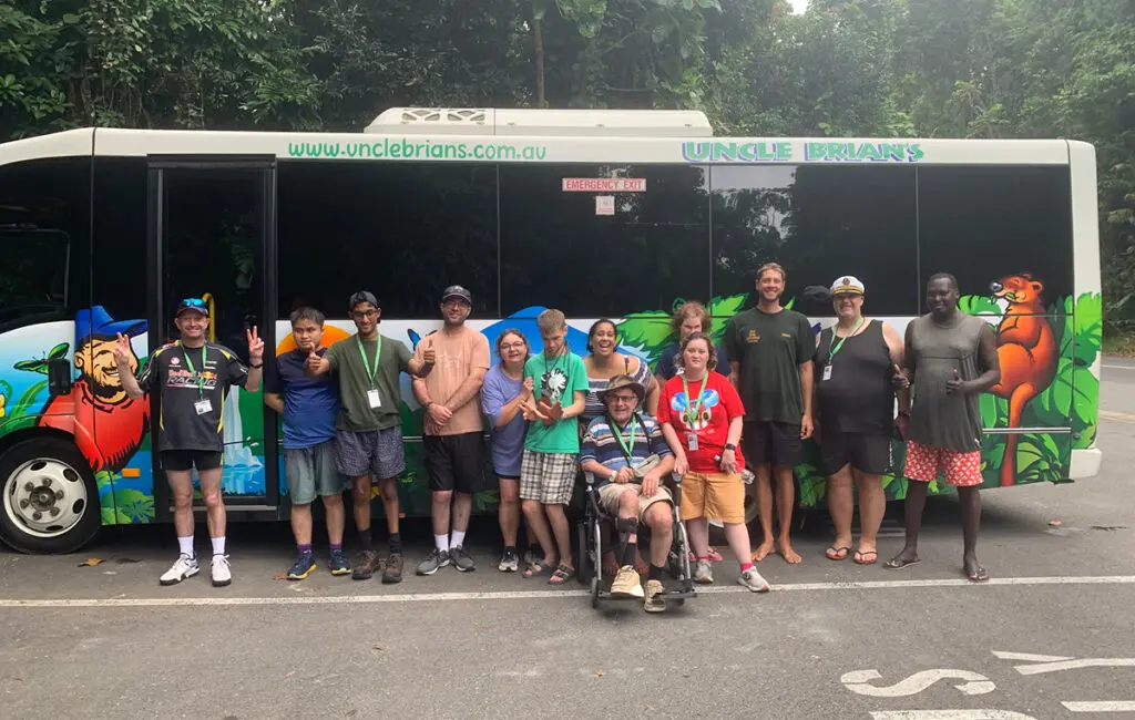 Group poses in front of an 'Uncle Brian's' tour bus, one person using a wheelchair, surrounded by rainforest.