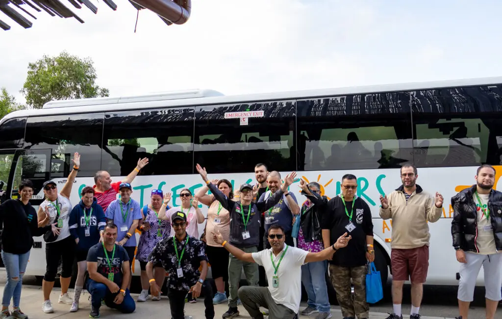 Group wearing lanyards waves and poses in front of a coach bus with painted sightseeing tours artwork.