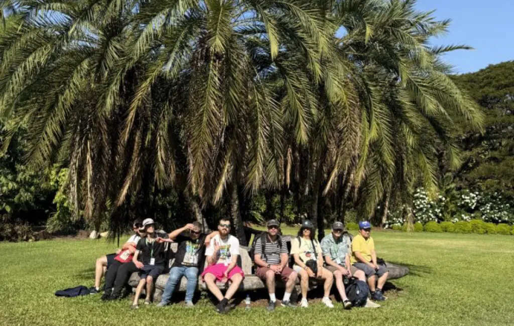 Group of eight people sit on a log bench under a large palm tree in a garden.