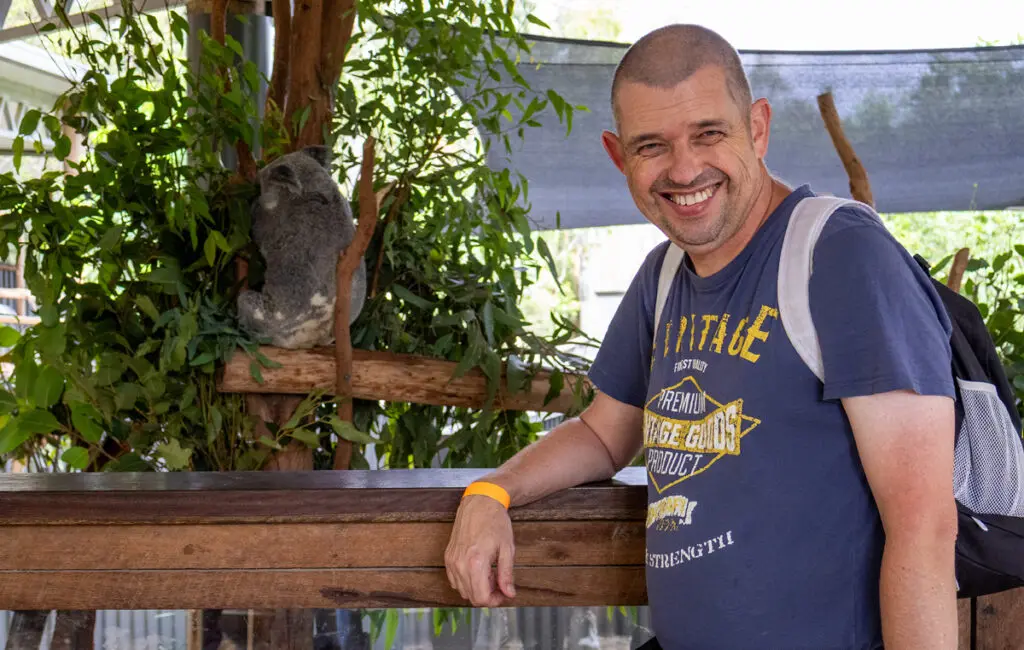 Man in a blue T-shirt smiles at a wildlife enclosure, with a koala resting on a branch behind him.