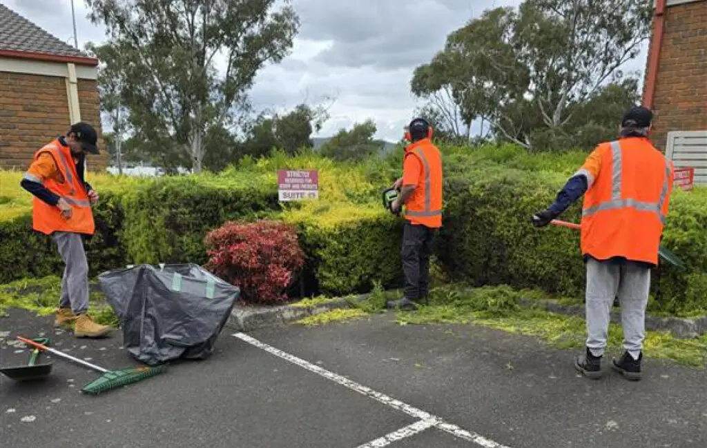 Three workers in orange hi-vis vests trim hedges in a car park, with a garden waste bag and rake nearby.