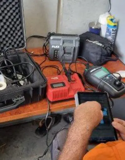 Man in orange shirt holds a tablet beside a workbench with battery chargers, a meter, and a tool case.