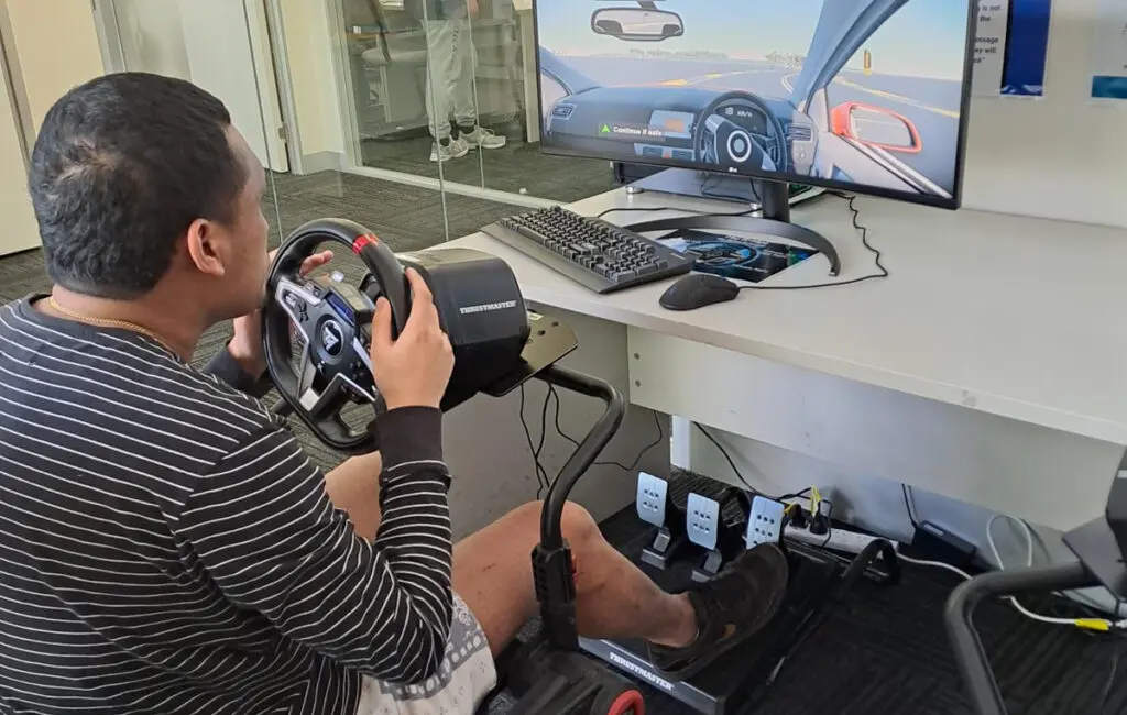 Man using a driving simulator with steering wheel and pedals, viewing a road scene on a monitor.