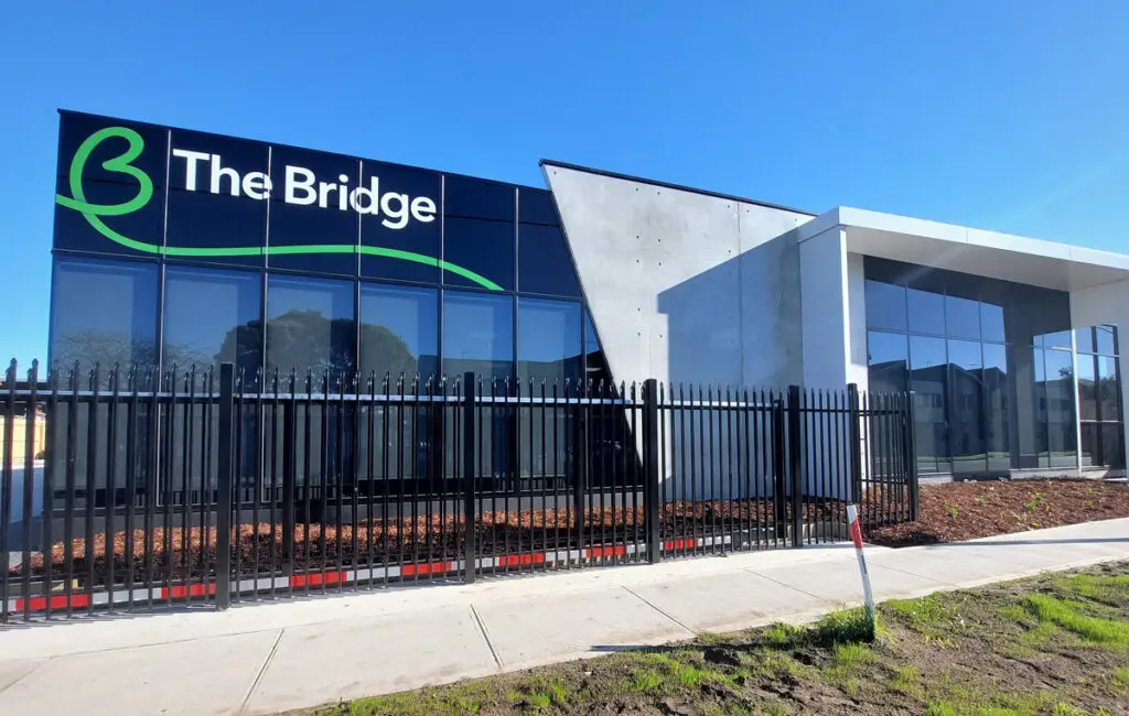 Modern building with 'The Bridge' sign and logo, glass windows, and a black steel perimeter fence.