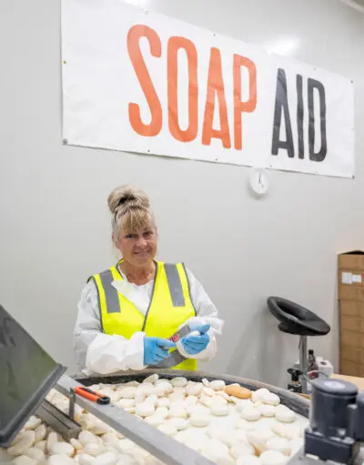 Woman in hi-vis vest and gloves sorting soap bars at a table, under a Soap Aid banner.