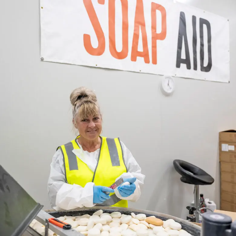 Woman in hi-vis vest and gloves sorting soap bars at a table, under a Soap Aid banner.