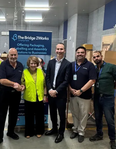 Five people stand together in a warehouse beside a Bridge 2Works Employment banner and boxes.