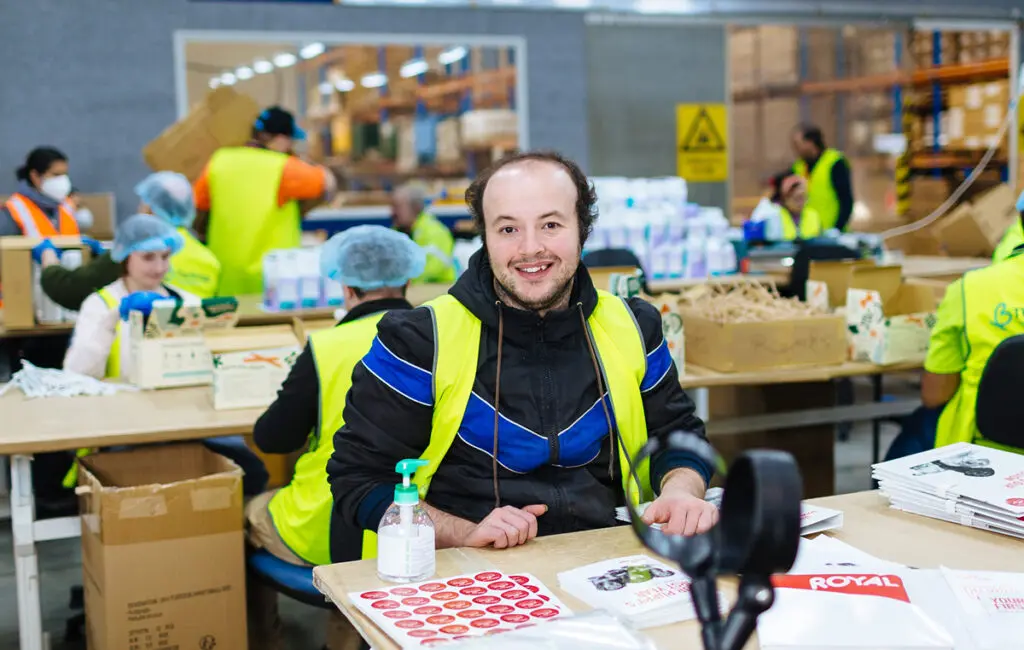 Man in high-vis vest smiles at a packing table in a warehouse, colleagues working behind him.