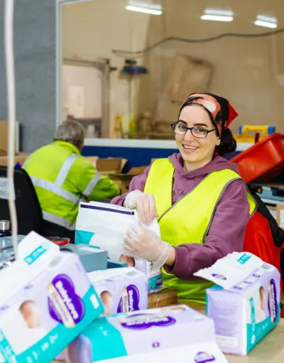 Woman in hi-vis vest and gloves smiling while packing nappies at a warehouse table, colleague working behind her