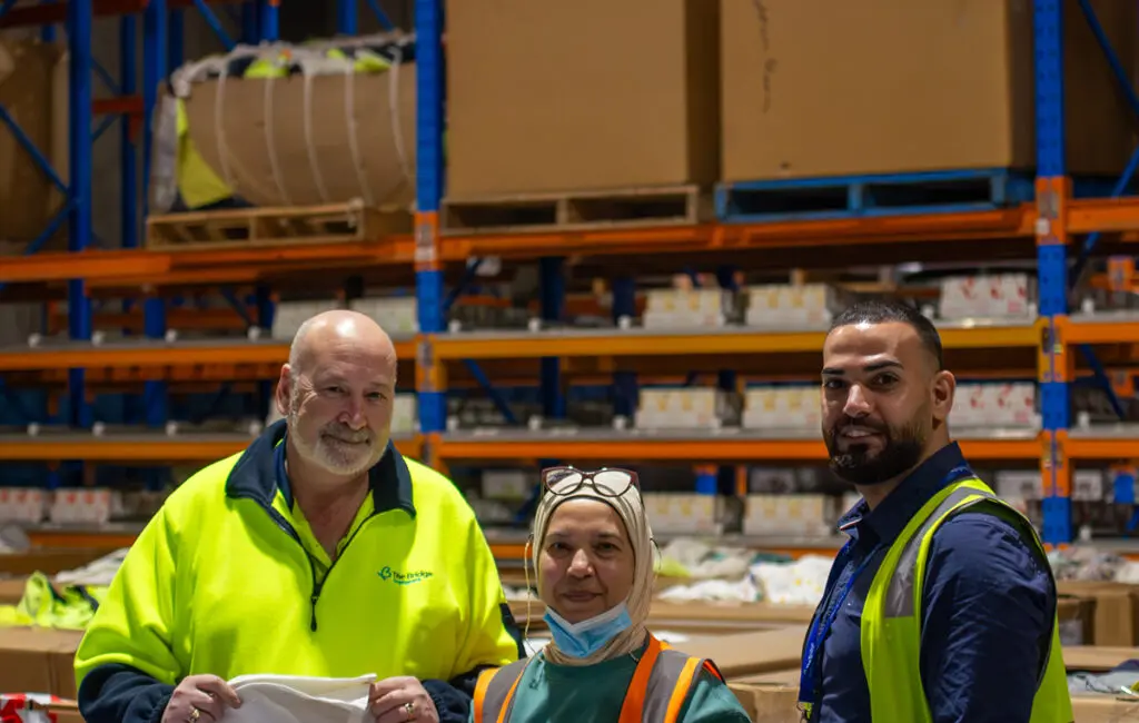 Three people in hi-vis vests stand in a warehouse aisle with pallet racking and boxes behind them.