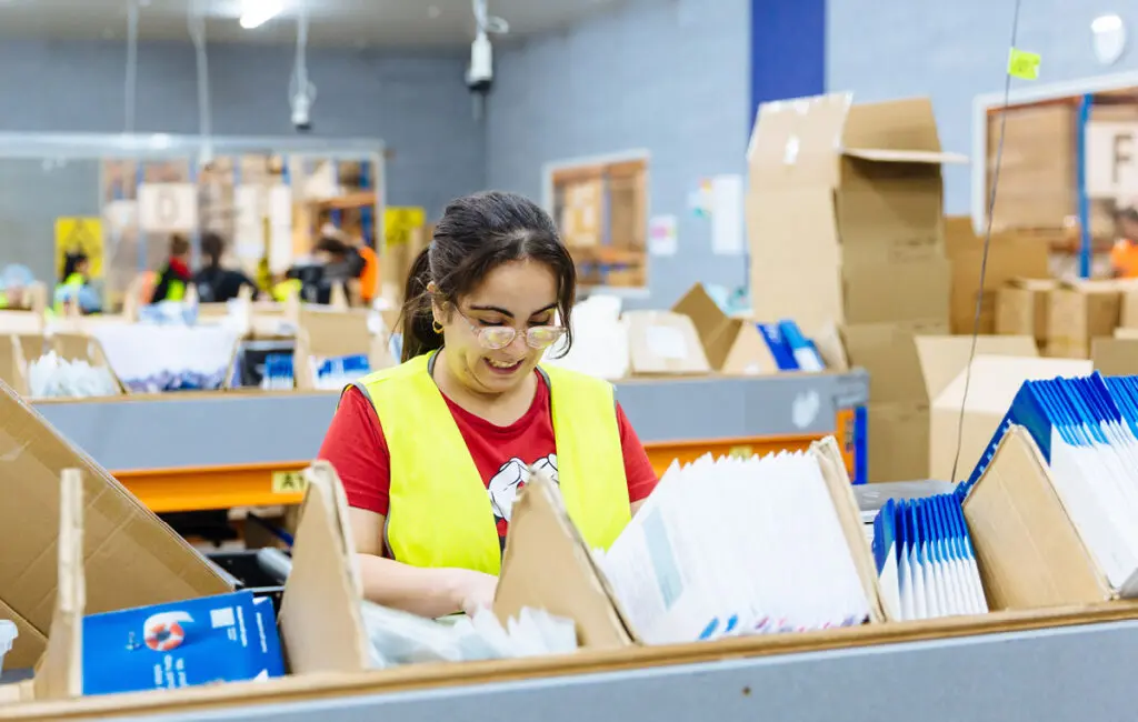Woman in glasses and a hi-vis vest sorts papers into cardboard boxes in a warehouse.