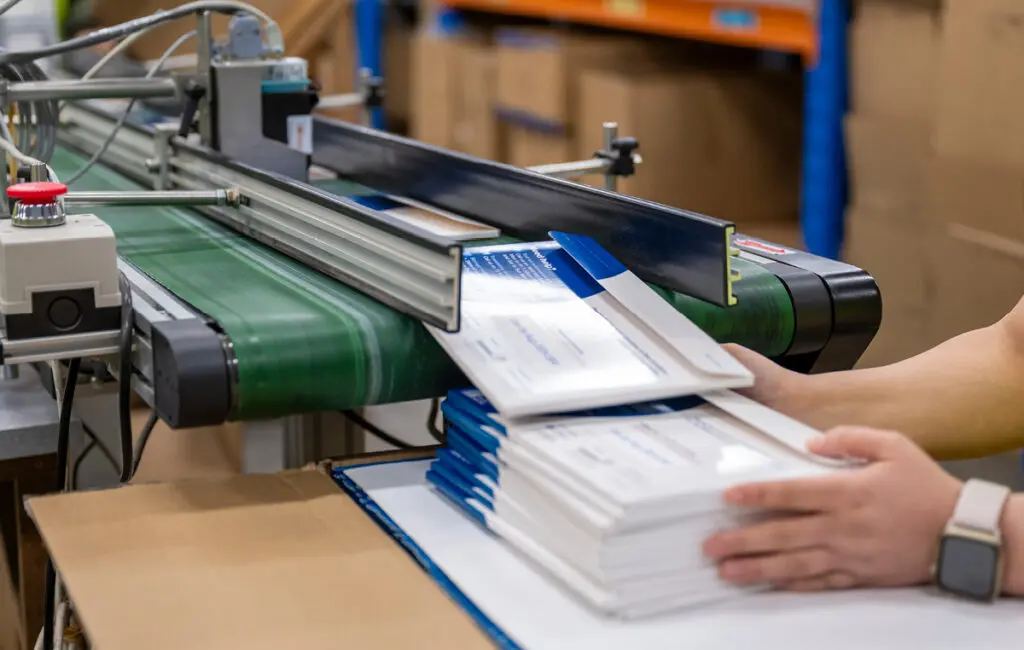 Hands stacking printed mailers as they come off a conveyor belt in a mailing facility.