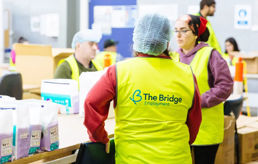 Workers in hi-vis vests reading "The Bridge Employment" and hairnets at a packing line with product bags.
