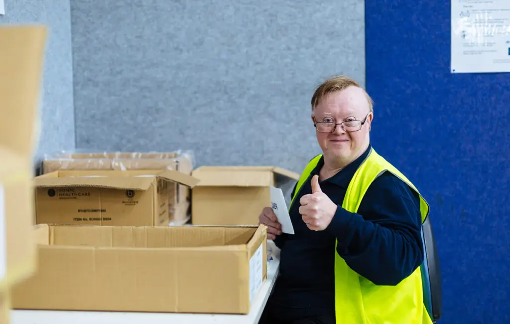 Man wearing glasses and a hi-vis vest gives a thumbs up beside a workbench with cardboard boxes.