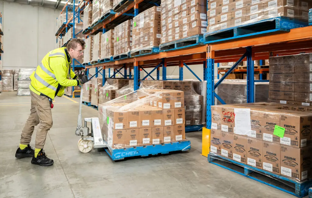 Warehouse worker in high-vis vest uses a pallet jack to move boxes among tall storage racks.