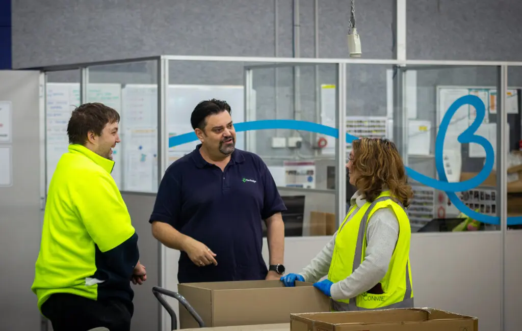 Two men and a woman in hi-vis talk near cardboard boxes on a trolley in a warehouse office.