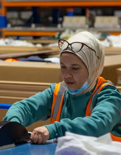 Woman in hijab and orange safety vest sorts items at a table in a warehouse.
