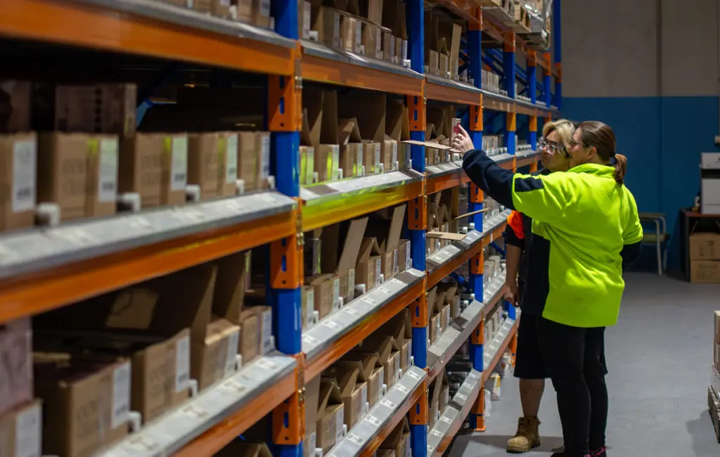Two workers in a warehouse select boxes from tall shelving stocked with cardboard cartons.