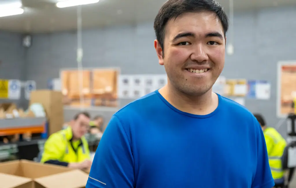 Man in a blue t-shirt smiles in a warehouse, with workers in high-vis vests and cardboard boxes behind him.