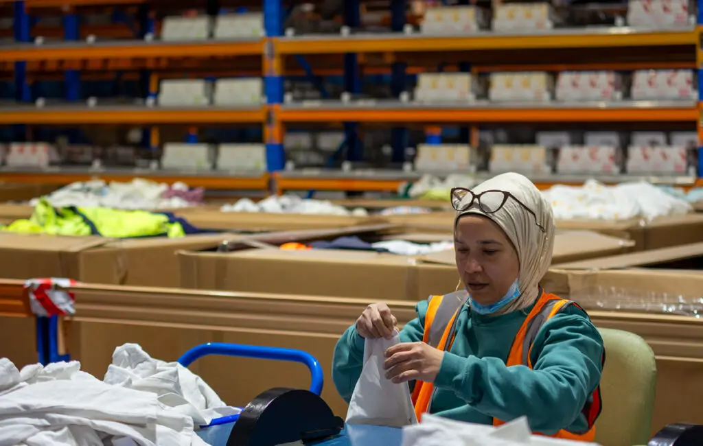 Woman in hijab and hi-vis vest packs folded clothing into bags at a warehouse sorting table.