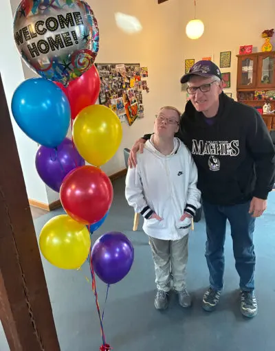 Two people stand indoors beside colourful balloons and a "Welcome Home!" balloon, one with an arm around the other's shoulder.