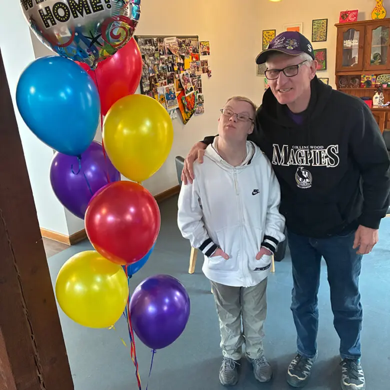 Two people stand indoors beside colourful balloons and a "Welcome Home!" balloon, one with an arm around the other's shoulder.
