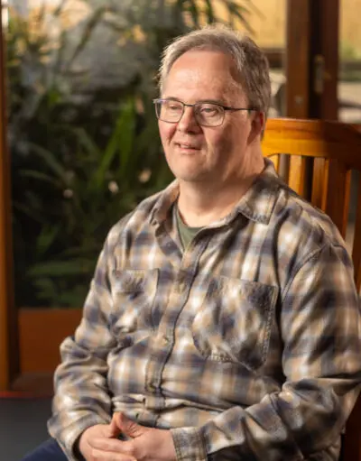 Man with glasses wearing a checked flannel shirt, sitting on a wooden chair indoors, plants visible behind him.