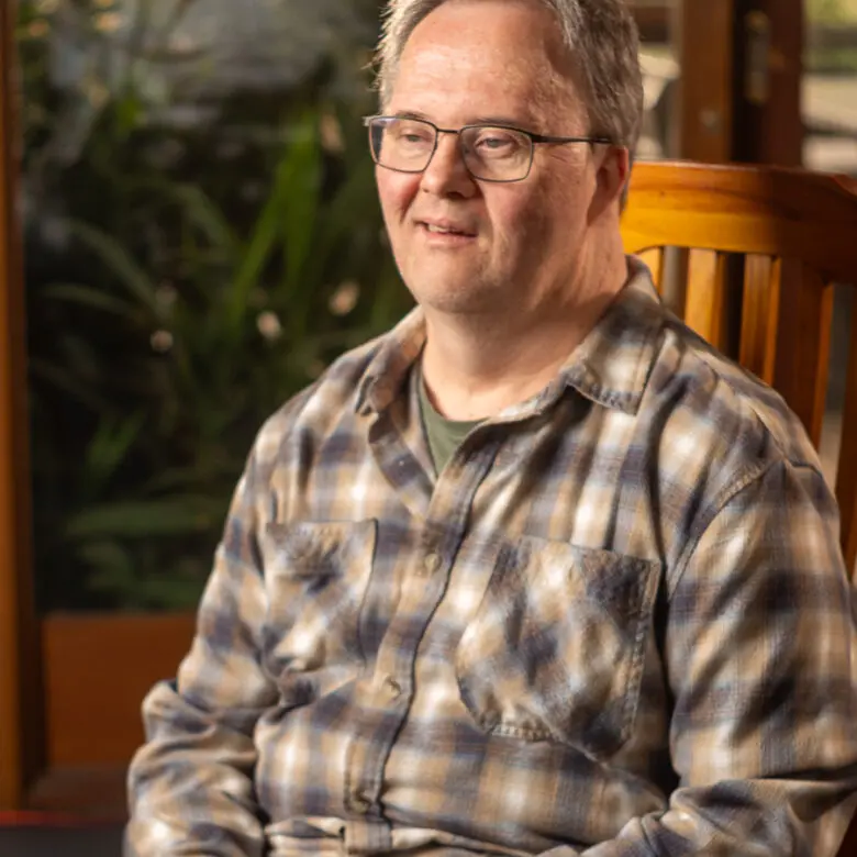 Man with glasses wearing a checked flannel shirt, sitting on a wooden chair indoors, plants visible behind him.