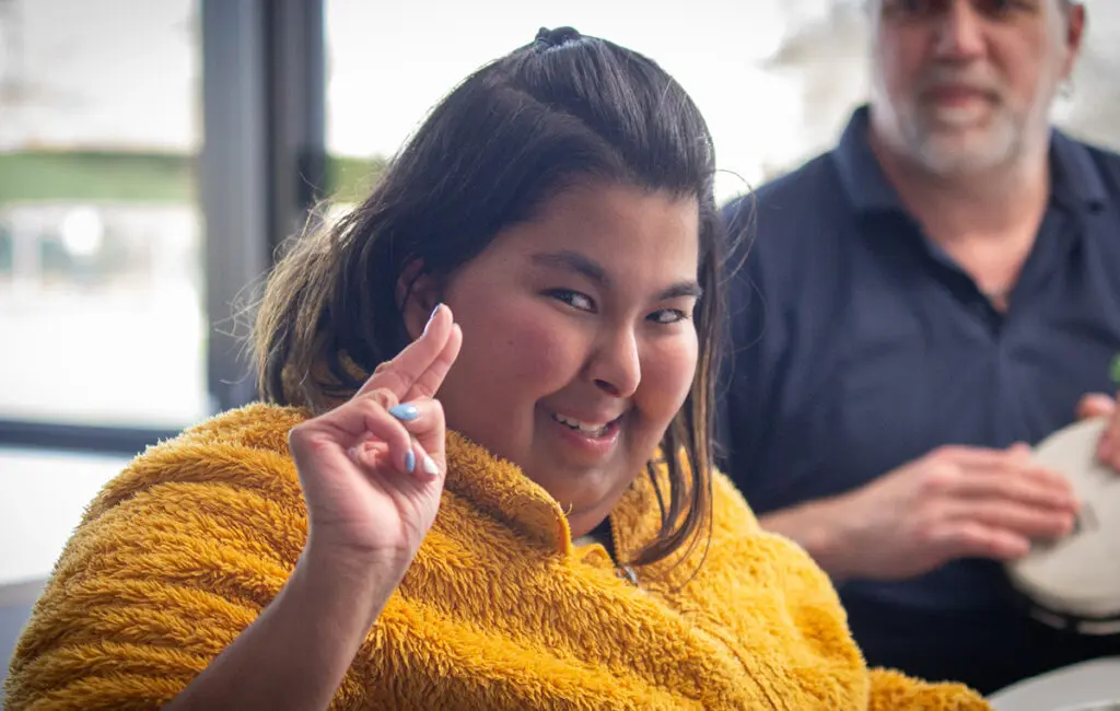 Woman in a yellow fleece wrap smiles, raising a hand; a man behind her holds a hand drum.