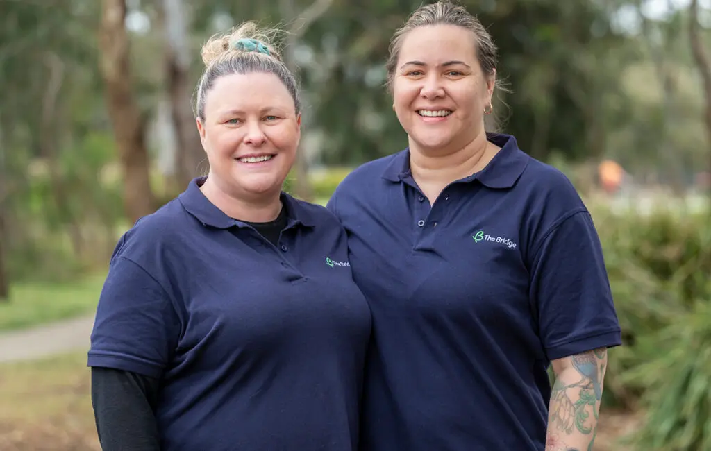 Two women in navy 'The Bridge' polo shirts smiling outdoors, trees in background.