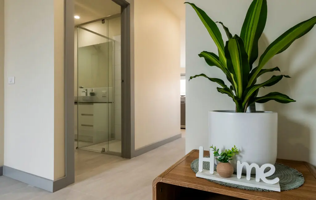 Hallway with potted plant on wooden table beside 'Home' sign, glass-walled bathroom visible through doorway.