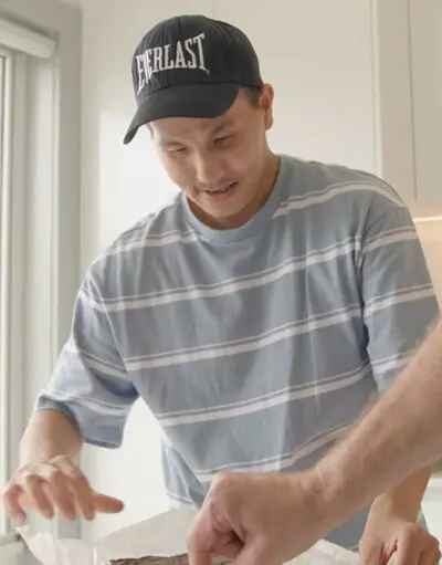 Man in an Everlast cap and striped shirt smiling while reaching toward a foil-lined tray in a kitchen.