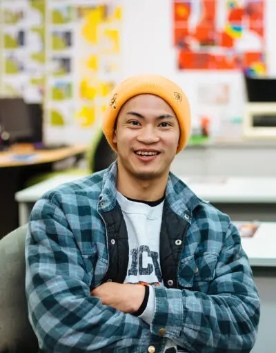 Man in an orange beanie and checked flannel shirt smiles with arms crossed in an office.
