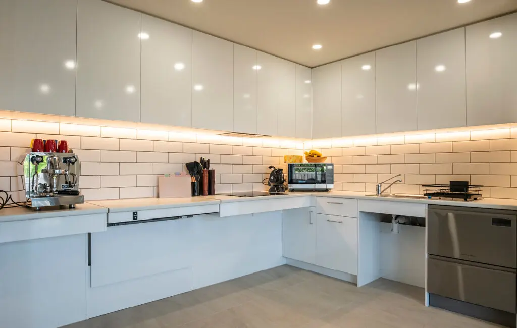 Accessible kitchen with lowered white benchtops, under-cabinet lighting, sink, cooktop and dishwasher drawers.