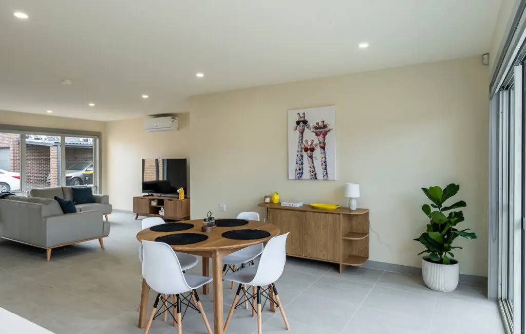 Open-plan living area with round dining table, white chairs, sideboard, giraffe artwork, sofa and TV unit.