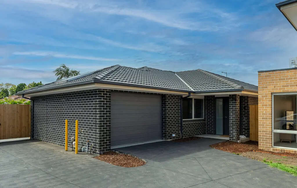Single-storey dark brick house with tiled roof, garage, front door and concrete driveway.
