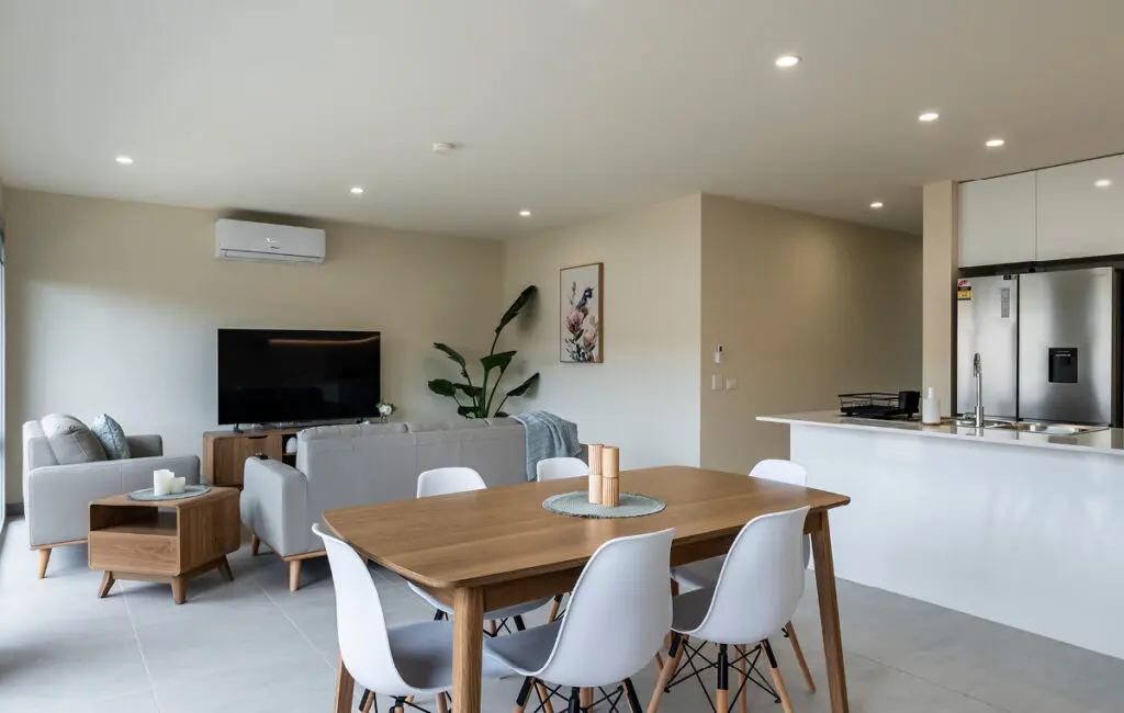 Open-plan living area with wooden dining table, white chairs, lounge, TV, and kitchen with fridge