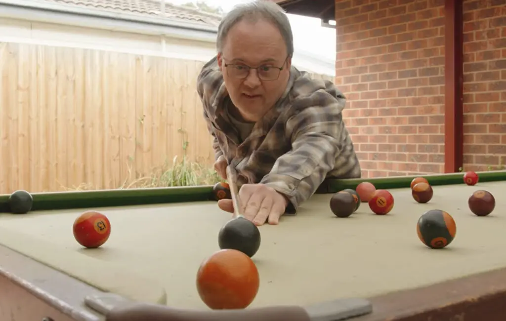 Man in a plaid shirt leaning over an outdoor pool table, aiming a cue at coloured balls.