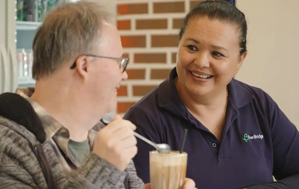 Man with an iced coffee smiles beside a support worker in a navy 'The Bridge' polo shirt.
