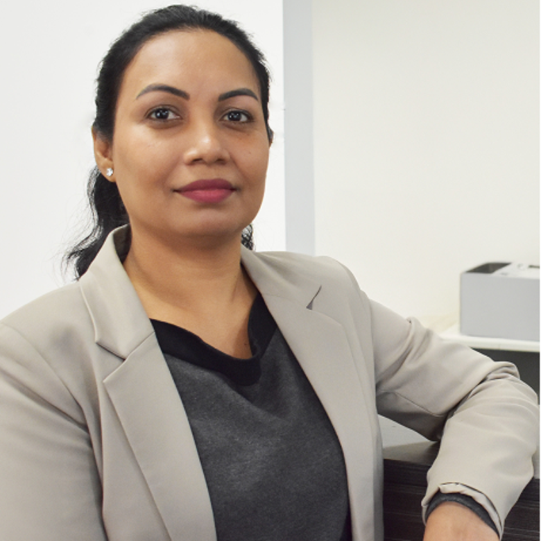 Woman with dark hair wearing a grey blazer over a dark top, seated in an office.