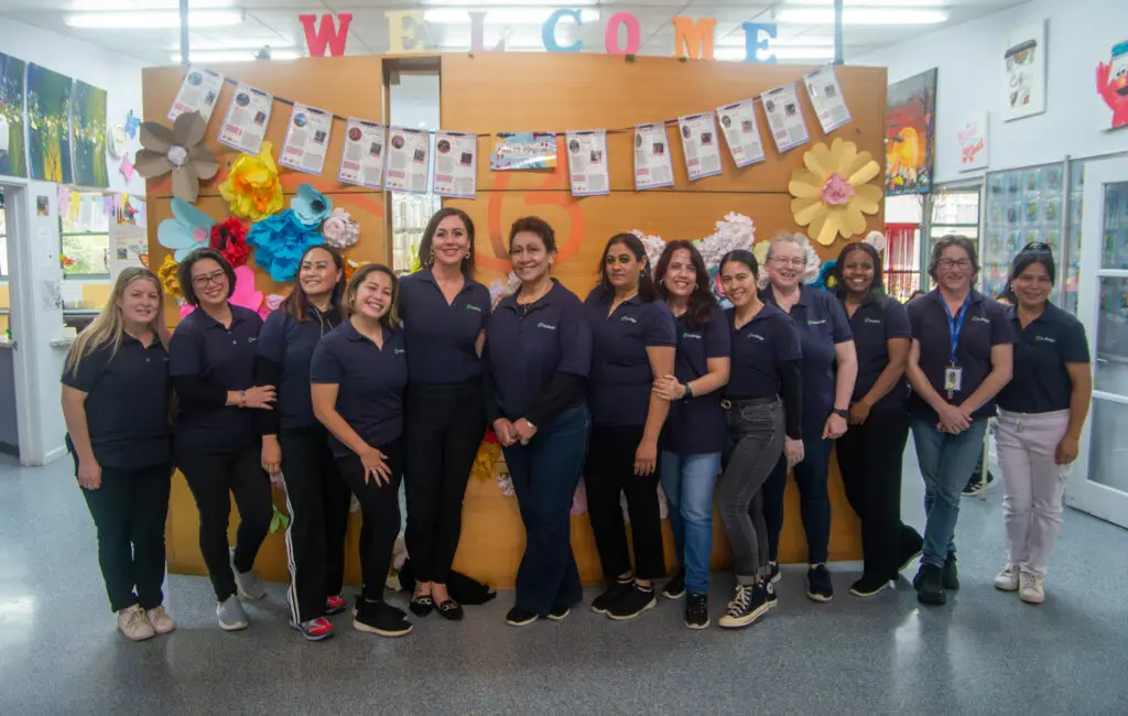 Group of women in matching navy polos stand together in a childcare centre, under a wooden welcome display.
