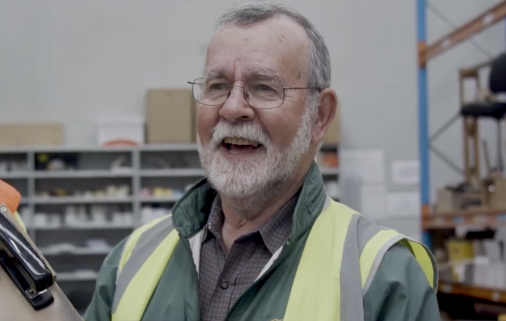 Man with glasses and grey beard wearing a hi-vis vest, speaking in a warehouse with shelving.