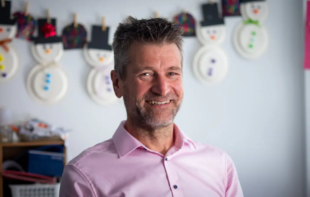 CEO smiling, standing in a classroom with paper-plate snowman crafts hanging behind him.