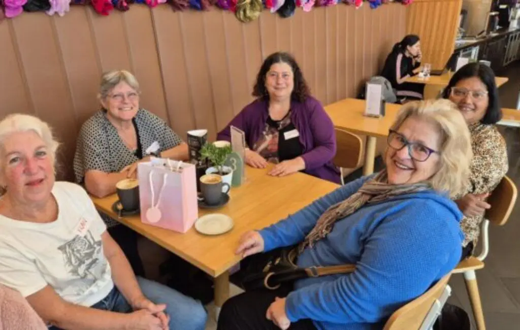 Five women sit around a café table with coffees and a pink gift bag, smiling at the camera.