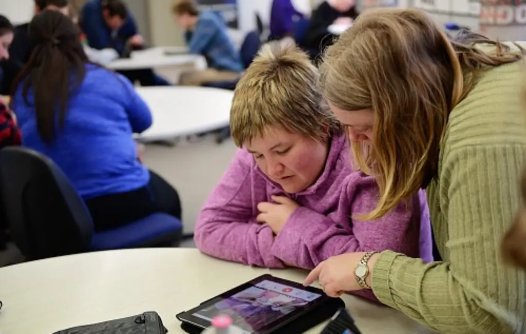 Woman in pink leaning on a table while another woman points at a tablet screen beside her.