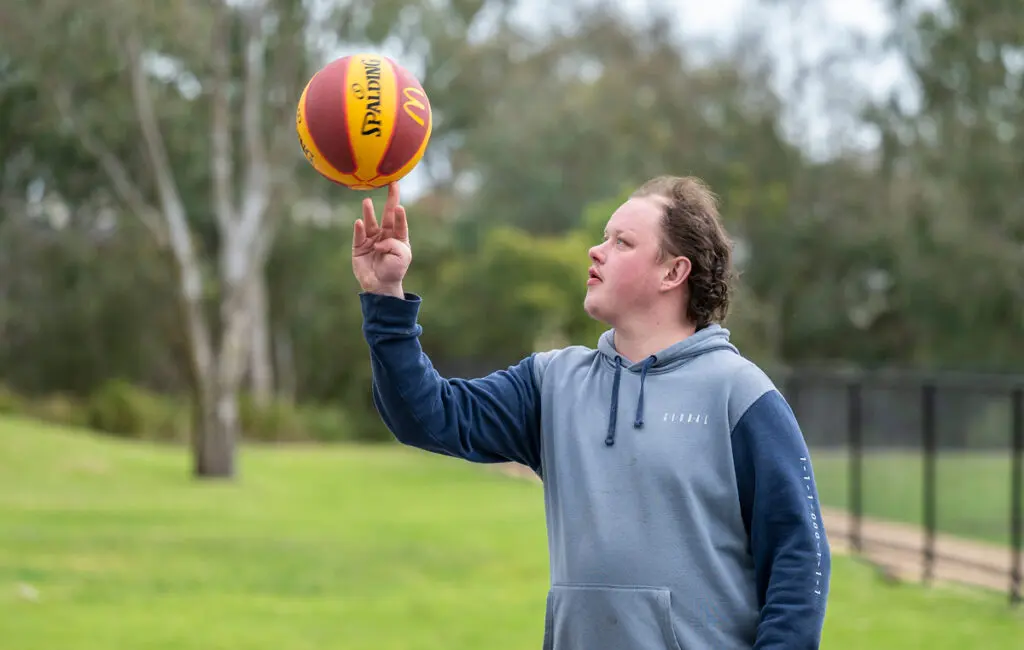 Young man in a hoodie balances a basketball on his fingertip in a grassy park.